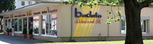 Möbelgeschäft mit großen Fenstern und gelbem Logo „bodeto Wohnen mit Stil“ an der Fassade.