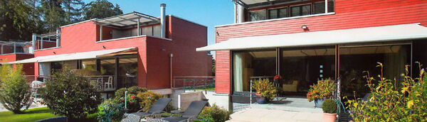 Moderne rote Holzhäuser mit großen Terrassen und Sonnenschutz im gepflegten Garten.