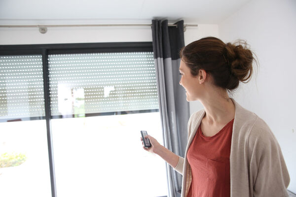 Frau bedient elektrische Rollläden mit Fernbedienung vor Fenster im modernen Wohnzimmer.